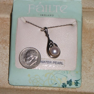 May include: A silver-toned necklace with a teardrop-shaped pendant featuring a white water pearl. The pendant has a Celtic knot design. The necklace is displayed on a light blue card with the text "Fáilte Ireland" and a dime for size comparison.