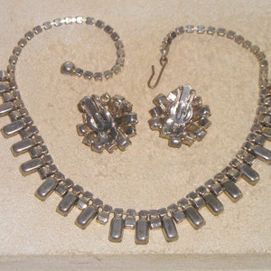 May include: A silver tone necklace and earrings set with clear rhinestones. The necklace has a long, rectangular design with a row of stones hanging down from the center. The earrings are clip-on and have a similar design to the necklace.