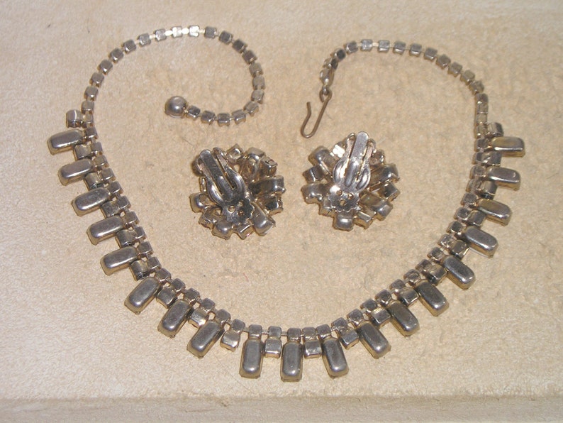 May include: A silver tone necklace and earrings set with clear rhinestones. The necklace has a long, rectangular design with a row of stones hanging down from the center. The earrings are clip-on and have a similar design to the necklace.