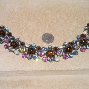Puede incluir: Un brazalete de metal con piedras de strass multicolores, con piedras marrones, azules, rosas y blancas. El brazalete tiene un diseño delicado e intrincado.