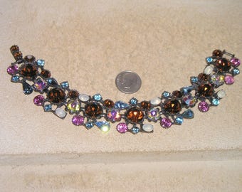 May include: A multi-coloured rhinestone bracelet featuring brown, blue, pink and white stones. The bracelet is made of metal and has a delicate, intricate design.