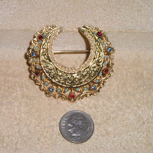 Puede incluir: Broche dorada en forma de media luna con intrincados detalles de filigrana y acentos de joyas. El broche presenta piedras rojas, azules y turquesas, y peque&ntilde;as perlas blancas. Se incluye una moneda como referencia de tama&ntilde;o.