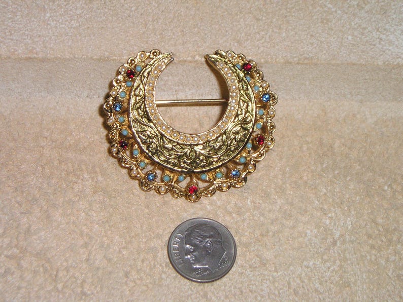 Puede incluir: Broche dorada en forma de media luna con intrincados detalles de filigrana y acentos de joyas. El broche presenta piedras rojas, azules y turquesas, y peque&ntilde;as perlas blancas. Se incluye una moneda como referencia de tama&ntilde;o.
