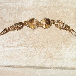 Puede incluir: Un brazalete dorado con dos piezas redondas, ornamentadas y decorativas en el centro. El brazalete tiene un cierre en cada extremo.