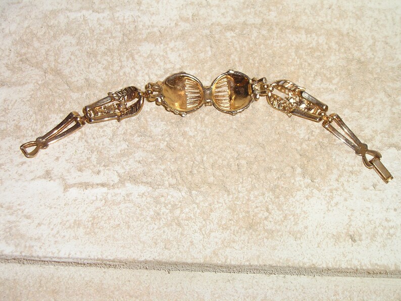 Puede incluir: Un brazalete dorado con dos piezas redondas, ornamentadas y decorativas en el centro. El brazalete tiene un cierre en cada extremo.