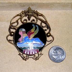 May include: A gold-toned brooch with an ornate design features a black enamel roundel with a colourful depiction of a winged sphinx. The sphinx is depicted in a blue, red, yellow, and green colour scheme against a black background. The brooch is approximately 5 cm in diameter.