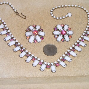 May include: A white and pink rhinestone necklace and earrings set. The necklace has a long, rectangular design with a row of white rhinestones and a row of pink rhinestones. The earrings are small and round, with a similar design to the necklace.