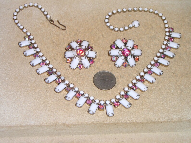 May include: A white and pink rhinestone necklace and earrings set. The necklace has a long, rectangular design with a row of white rhinestones and a row of pink rhinestones. The earrings are small and round, with a similar design to the necklace.