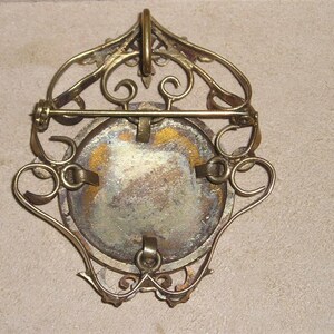 May include: A gold-toned metal brooch with a filigree design and a round, blank cameo. The brooch has a pin and catch closure on the back.