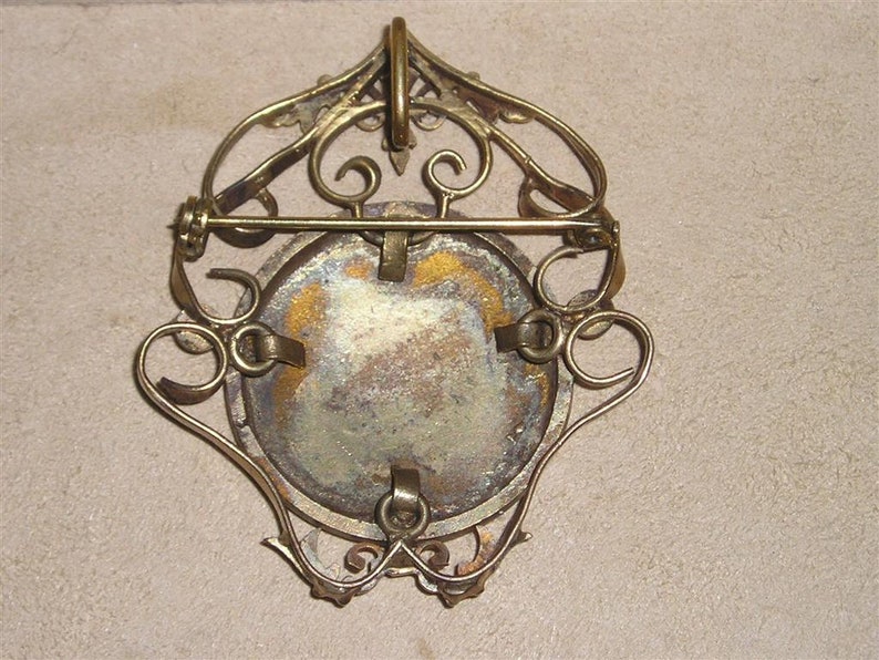 May include: A gold-toned metal brooch with a filigree design and a round, blank cameo. The brooch has a pin and catch closure on the back.