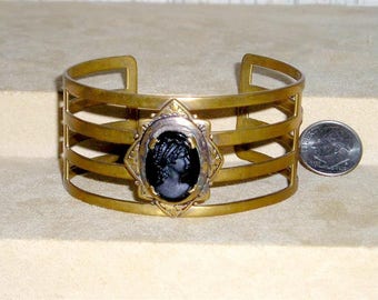 May include: A gold-toned metal bracelet with a black cameo featuring a woman's profile. The bracelet has a multi-layered design with five horizontal bars.