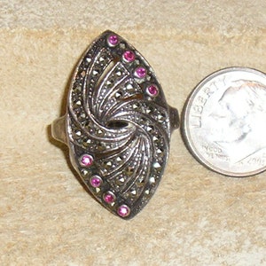 May include: A silver ring with a marquise-shaped setting featuring a swirling design of marcasite stones and pink accents.