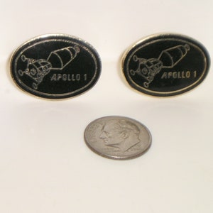 May include: Two gold-colored oval pins with black enamel. Each pin features a black and white illustration of the Apollo 1 spacecraft and the text "APOLLO 1".