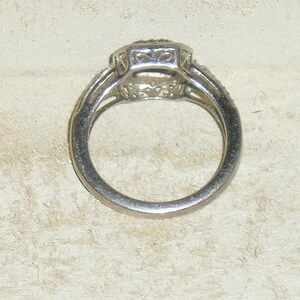 May include: A silver ring with a square-shaped center stone surrounded by smaller stones. The ring has a delicate design and is set on a white background.