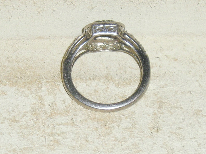 May include: A silver ring with a square-shaped center stone surrounded by smaller stones. The ring has a delicate design and is set on a white background.