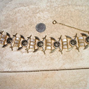 May include: A vintage gold-tone bracelet with a decorative design. The bracelet features alternating sections of white rectangular elements and ornate gold-tone accents. A dime is included for size reference. The bracelet has a chain and clasp closure.