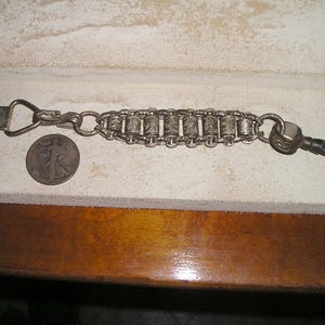 May include: A silver-toned metal watch chain featuring a series of seven cylindrical links with engraved patterns, connected by small rings. The chain includes a flat rectangular clip on one end and a swivel hook on the other, positioned next to a coin.