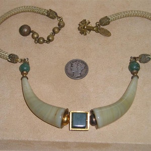 May include: Vintage necklace featuring a cream-colored woven cord and brass-toned metal accents. The necklace has two curved, light green horn-shaped pendants with a square, teal-colored center stone. A green bead and a silver coin are also visible.