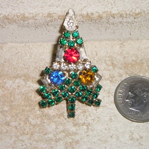 Puede incluir: Un broche de árbol de Navidad de cristal verde con cristales rojos, azules y amarillos en el centro.