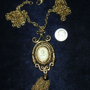 May include: A gold chain necklace with a cameo pendant featuring a white rose design. The pendant is surrounded by a gold frame and has a tassel made of gold chain.