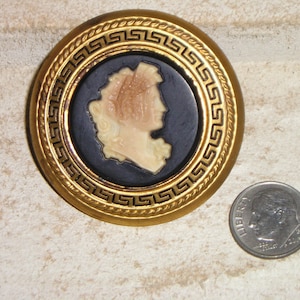 May include: A gold-toned brooch with a black and white cameo of a woman's profile. The brooch is decorated with a Greek key pattern.