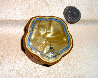 May include: A small, round, gold-colored trinket box with a blue and white design. The box has a wavy, scalloped edge and a raised design on the lid.