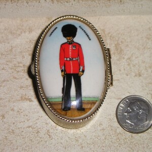 May include: A small oval-shaped metal trinket box with a hinged lid. The lid features a colorful image of a London Guardsman in full uniform. The text "LONDON GUARDSMAN" is printed around the image.