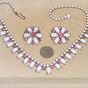 May include: A white and pink rhinestone necklace and earrings set. The necklace has a long, rectangular pendant design with a row of smaller rhinestones on each side. The earrings are a matching flower design.