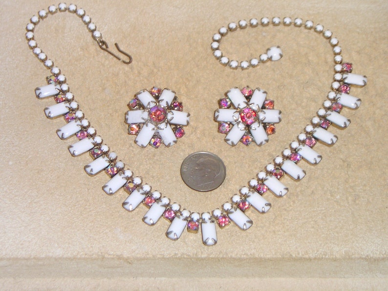 May include: A white and pink rhinestone necklace and earrings set. The necklace has a long, rectangular pendant design with a row of smaller rhinestones on each side. The earrings are a matching flower design.