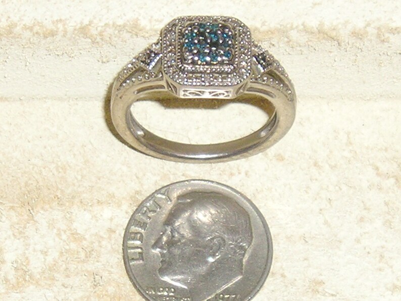 May include: A silver ring with a square-shaped blue diamond surrounded by smaller white diamonds. The ring is next to a US dime for size comparison.