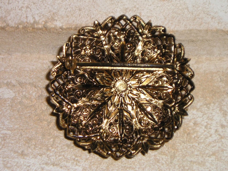 K&ouml;nnte beinhalten: Verzierte goldfarbene Brosche mit detailliertem Blumen- und Filigran-Design. Die R&uuml;ckseite hat einen Nadelverschluss f&uuml;r sicheren Halt. Die Brosche hat eine Vintage-&Auml;sthetik, perfekt, um jedem Outfit einen Hauch von Eleganz zu verleihen.