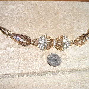 Puede incluir: Un brazalete de tono dorado con dos grandes grupos de diamantes de imitaci&oacute;n redondos y transparentes. El brazalete tiene un dise&ntilde;o de hoja a cada lado de los grupos.