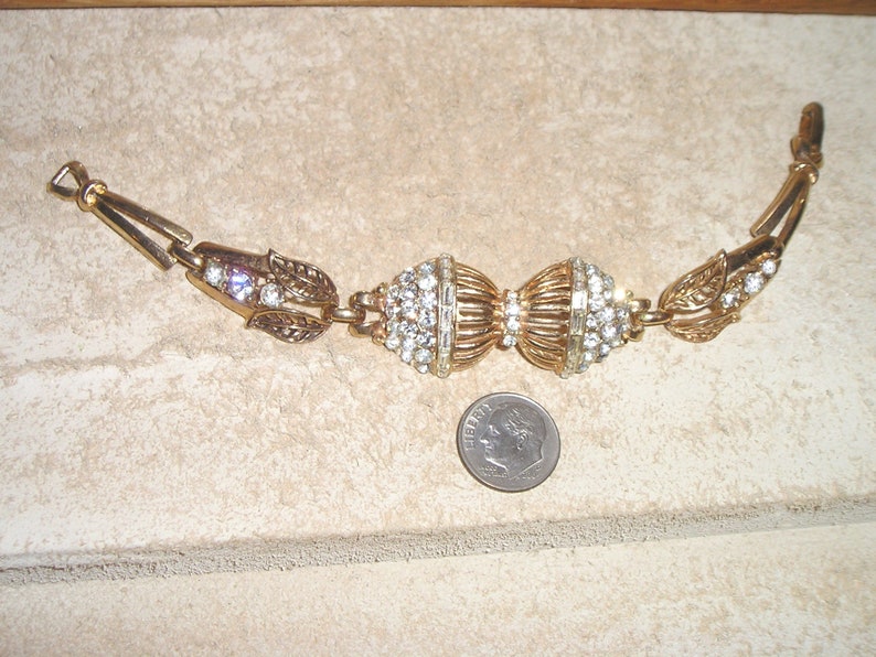 Puede incluir: Un brazalete de tono dorado con dos grandes grupos de diamantes de imitaci&oacute;n redondos y transparentes. El brazalete tiene un dise&ntilde;o de hoja a cada lado de los grupos.