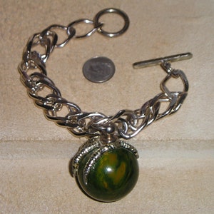 May include: A silver chain bracelet with a green and gold ball charm. The ball is attached to the chain with a silver claw design.