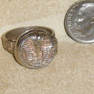 May include: Silver-toned ring with a detailed, raised design of an Egyptian pharaoh's head. The ring is next to a U.S. dime coin for size comparison. The ring's band has a repeating pattern. The ring is likely made of metal.