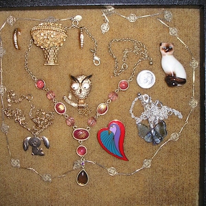 May include: A collection of vintage jewelry displayed in a black velvet-lined box. The jewelry includes a gold owl brooch, a gold heart-shaped brooch, a gold bird brooch, a gold necklace with pink and red beads, a silver necklace with a silver bird pendant, and a silver necklace with a silver heart pendant.