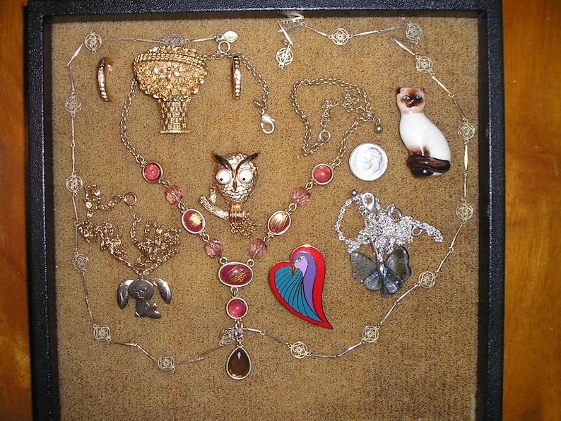 May include: A collection of vintage jewelry displayed in a black velvet-lined box. The jewelry includes a gold owl brooch, a gold heart-shaped brooch, a gold bird brooch, a gold necklace with pink and red beads, a silver necklace with a silver bird pendant, and a silver necklace with a silver heart pendant.