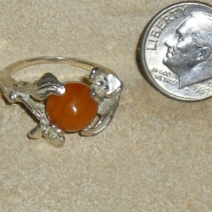 May include: A silver ring with a large orange gemstone set in a floral design. The ring is shown next to a US dime for size comparison.