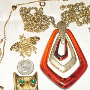 May include: A gold chain necklace with a large amber pendant featuring three interlocking geometric shapes. The pendant is accented with gold trim. The necklace is accompanied by a gold bee brooch and a gold owl brooch.