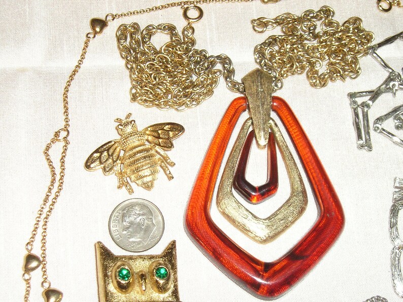 May include: A gold chain necklace with a large amber pendant featuring three interlocking geometric shapes. The pendant is accented with gold trim. The necklace is accompanied by a gold bee brooch and a gold owl brooch.
