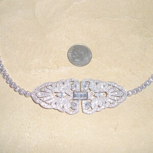 May include: A silver bracelet with a large, ornate, diamond-shaped center piece and a row of smaller diamonds around the edge. The bracelet is on a light brown surface.