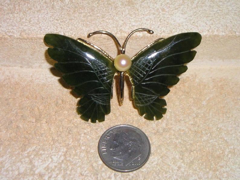May include: A dark green jade butterfly brooch with gold-coloured accents and a pearl centre. The butterfly's wings are intricately carved. A coin is placed next to the brooch for size comparison.