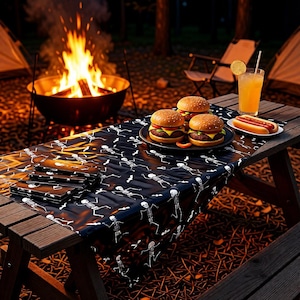 May include: A black plastic tablecloth featuring a repeating white skeleton pattern draped over a wooden picnic table. The table is set with three cheeseburgers on a black plate, a hot dog on a small white plate, and a glass of orange juice.