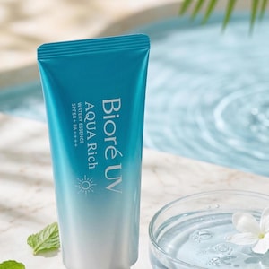 Puede incluir: Un tubo de protector solar Bioré UV Aqua Rich Watery Essence con SPF50+ PA++++. El tubo azul y blanco se encuentra sobre un fondo de agua, vegetación y un plato de vidrio con agua y una flor blanca.