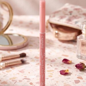 Puede incluir: Un lápiz delineador de ojos rosa CANMAKE Creamy Touch Liner Sheer (01). El producto de maquillaje está de pie sobre una superficie moteada, con brochas de maquillaje y un espejo compacto en el fondo. Pétalos de rosa secos están esparcidos alrededor del delineador.