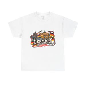 May include: A white short-sleeve t-shirt featuring a graphic print of German landmarks including the Cologne Cathedral and Brandenburg Gate, with the text "GERMANY", "DEUTSCHLAND", and "ARRIVED" alongside the German flag and a small airplane.