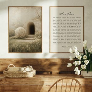 May include: Two framed art prints hang on a wall. One depicts a sepia-toned scene of a tomb with a large stone and a dark entrance. The other print has the text "He is Risen" with a passage below. A bouquet of white tulips sits on a wooden table.