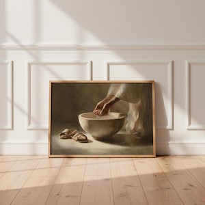 Puede incluir: Una impresión enmarcada que representa una escena religiosa. La obra de arte muestra manos sosteniendo agua sobre un cuenco de piedra. Sandalias descansan en el suelo frente al cuenco. La obra está enmarcada en un marco de madera marrón claro.