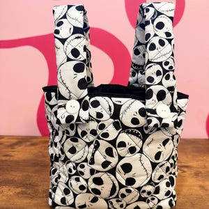 May include: A small tote bag with a black and white skull pattern. The bag has two handles and is secured with white buttons. The interior is black. The bag is made of a quilted fabric and sits on a wooden surface.
