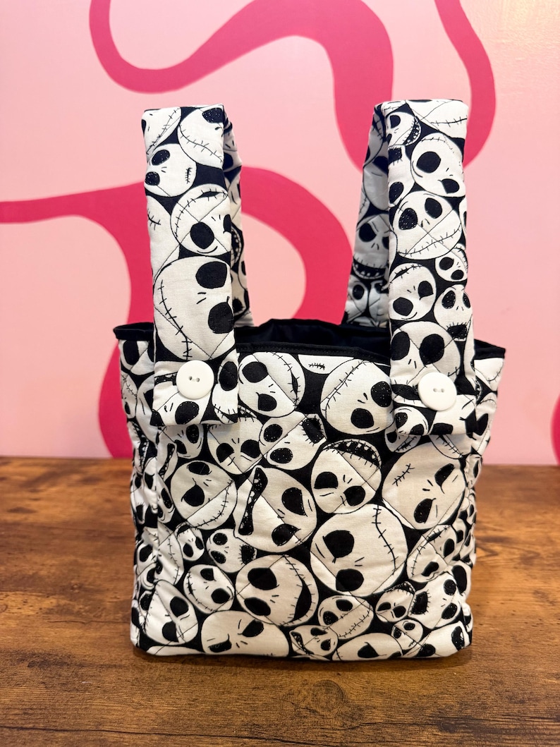 May include: A small tote bag with a black and white skull pattern. The bag has two handles and is secured with white buttons. The interior is black. The bag is made of a quilted fabric and sits on a wooden surface.